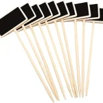 The image shows rustic chalkboard plant labels on stakes in a garden setting.